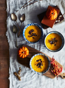 High angle close up of orange pumpkins and bowls of pumpkin soup on a table. High angle close up of orange pumpkins and bowls of pumpkin soup on a table.